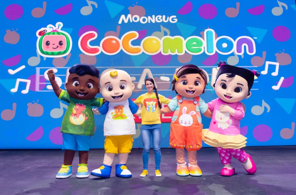 A group of four people in colorful CoComelon character costumes stand on stage with a smiling woman in a yellow shirt and jeans. They are in front of a bright blue backdrop with the Moonbug and CoComelon logos, music notes, and playful shapes. Everyone is posing with arms raised or linked, conveying a cheerful and energetic vibe.