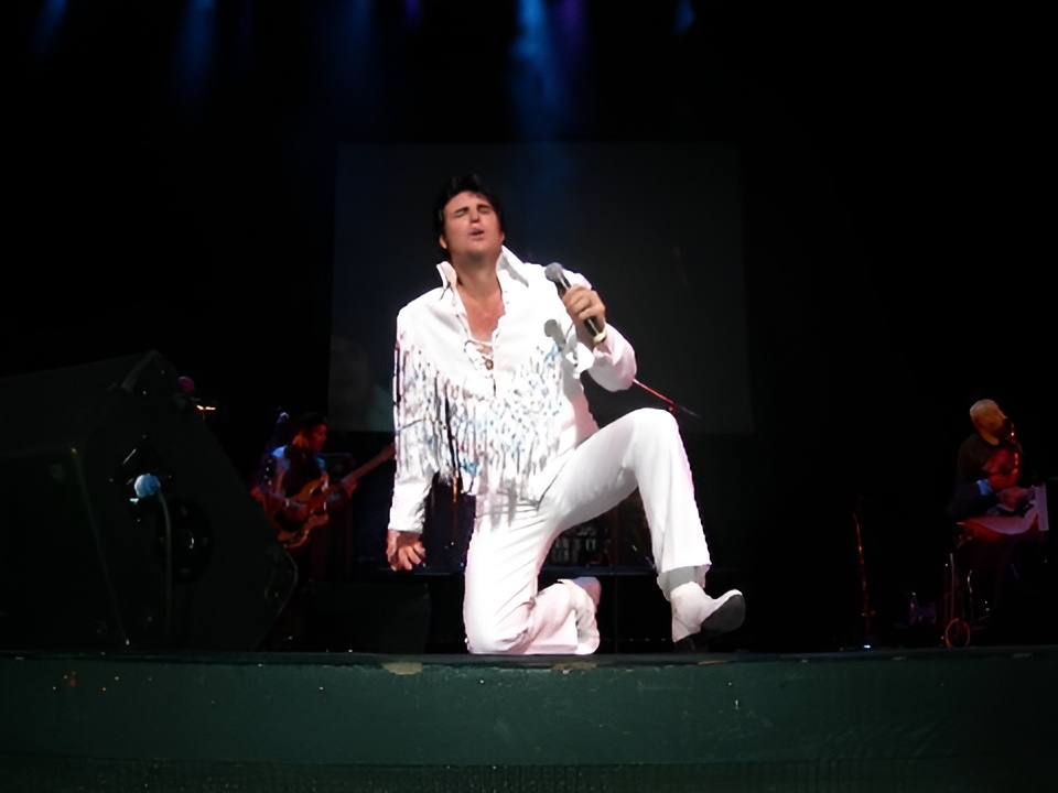 Chris MacDonald, who looks like Elvis, in a white fringed jumpsuit kneels on stage while singing into a microphone, with band members playing instruments in the background under dramatic stage lighting.
