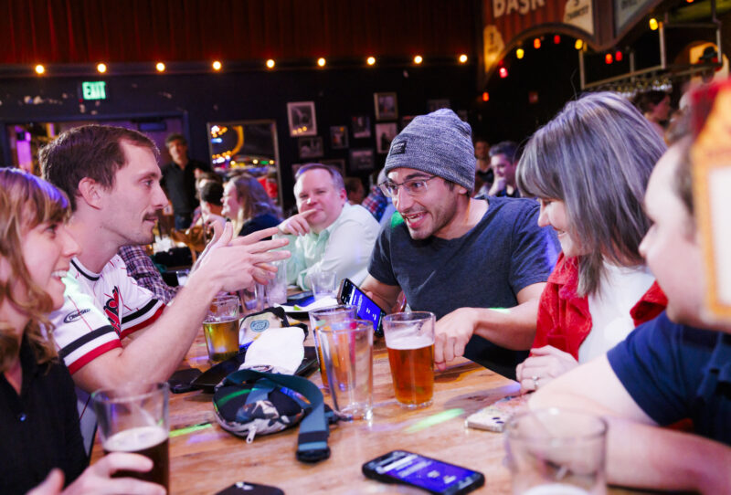 Group of people sitting around a wooden table in a bar, animatedly talking and laughing during a Jeopardy! Bar League trivia night. Several glasses of beer and smartphones are on the table. The atmosphere is lively with colorful lighting and decorations.