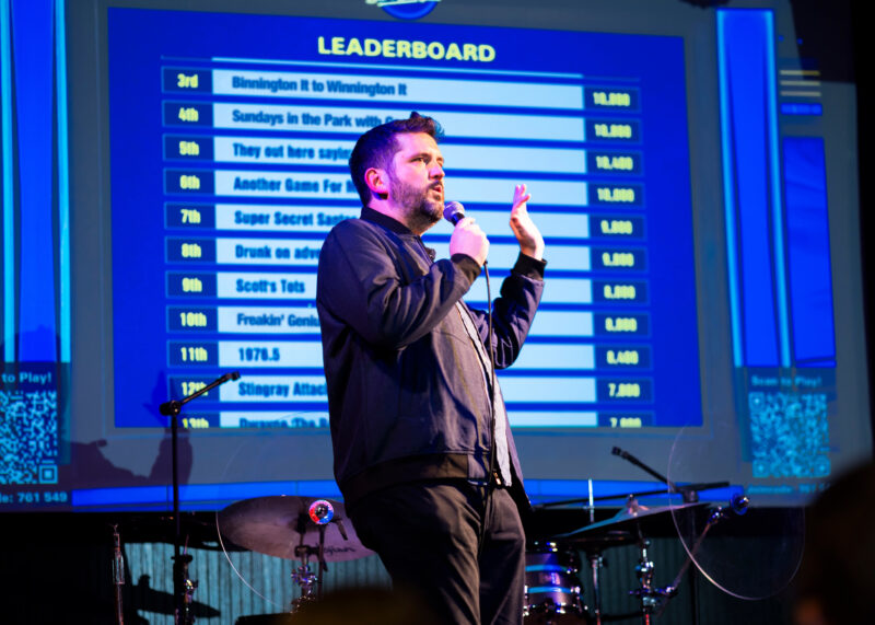 Man holding a microphone speaks on stage in front of a large projected Jeopardy! Bar League trivia leaderboard. He gestures with one hand, standing near a drum set with a blue and purple lighting setup.